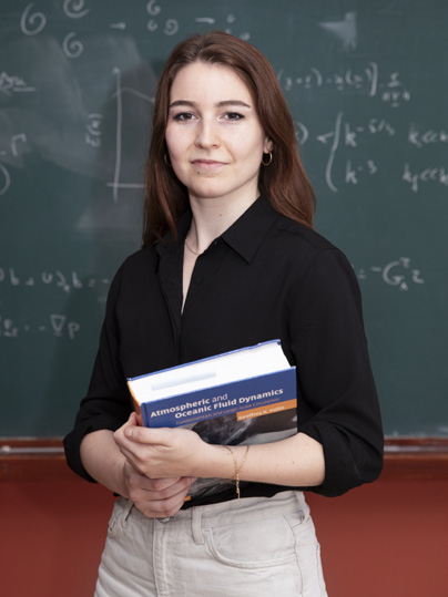 Portrait de Julie Meunier devant un tableau noir et portant un livre intitulé "Atmospheric et oceanic fluid dynamics"